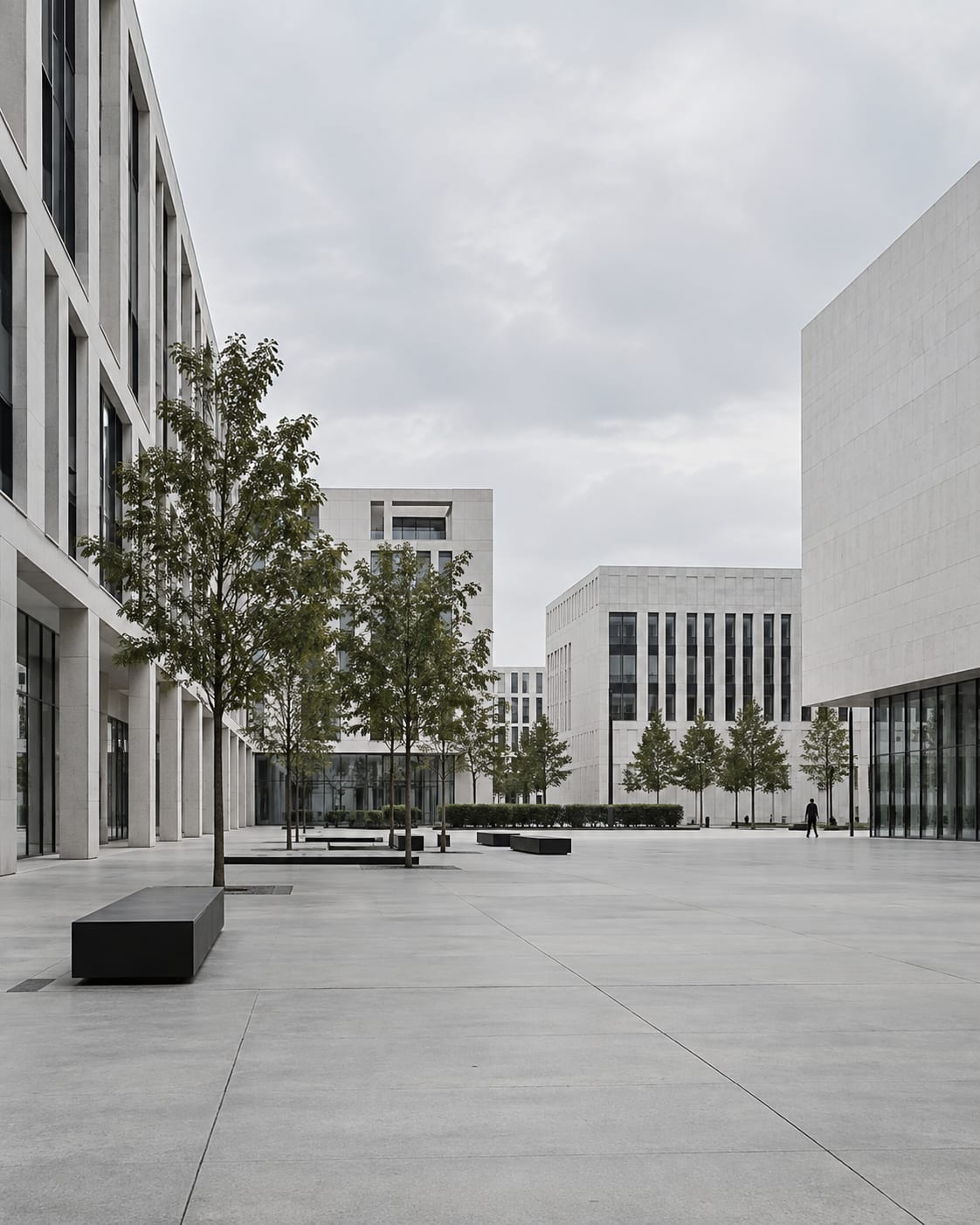Praça ampla de piso claro entre prédios de pedra, com árvores jovens, bancos baixos e uma pessoa caminhando ao fundo.